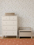 A white dresser and a matching storage bench sit against a wall adorned with Delicate Meadows Wallpaper Sample, showcasing its vibrant color and premium quality, with a woven basket on top. The floor features an orange rug with white polka dots.
