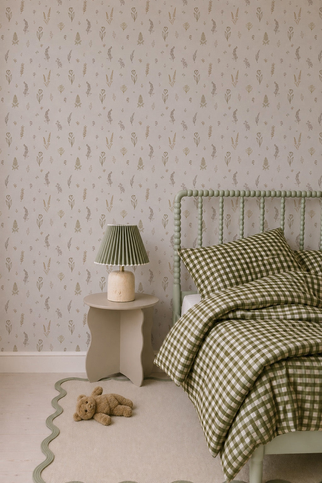 A cozy bedroom features a green checkered bed with matching pillow and blanket, complemented by the Delicate Meadows Wallpaper that adds charm and warmth to the space. A small round table holds a lamp with a green pleated shade, while a teddy bear rests on a beige rug bordered with wavy green edges.