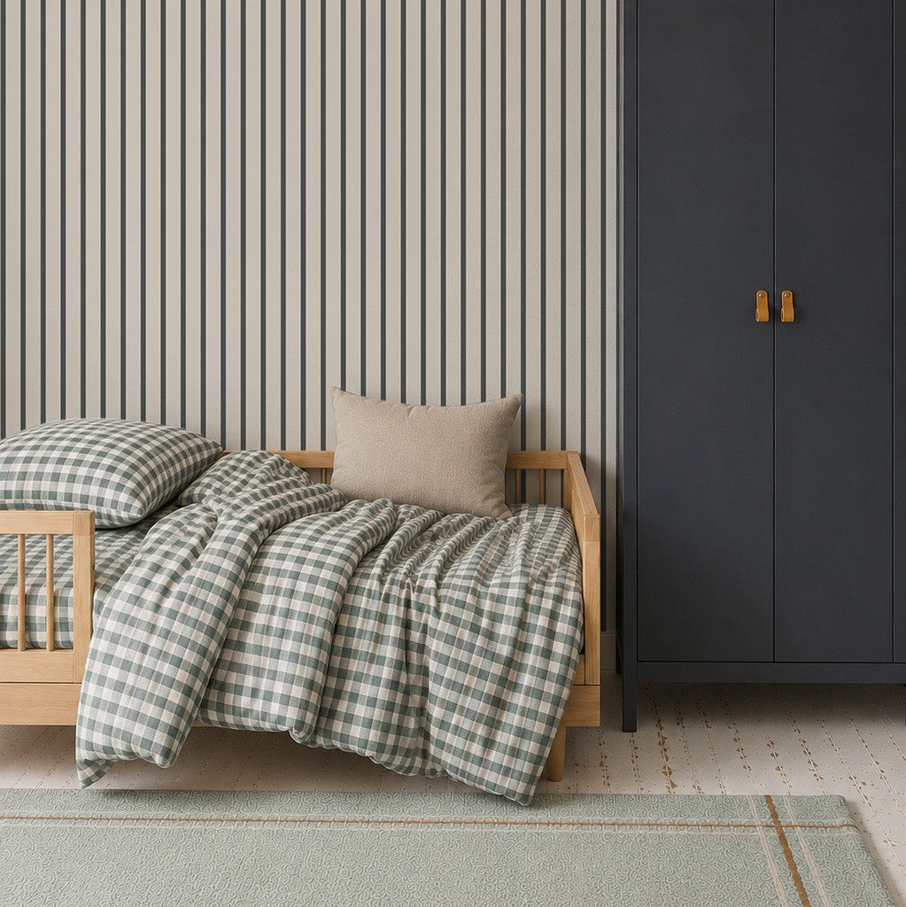 A cozy bedroom features a light wood toddler bed with checkered bedding and a beige pillow. A dark blue wardrobe with a basket on top stands nearby, set against Pinstripe Wallpaper Denim. A large light blue rug covers the floor.