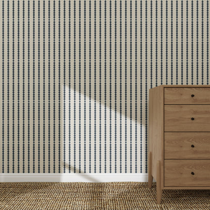 A wooden dresser stands on a woven rug beside a wall featuring the Little Abacus Stripe Denim Wallpaper sample. Sunlight casts a diagonal shadow, highlighting the wallpaper’s quality and vibrant blue striped pattern.
