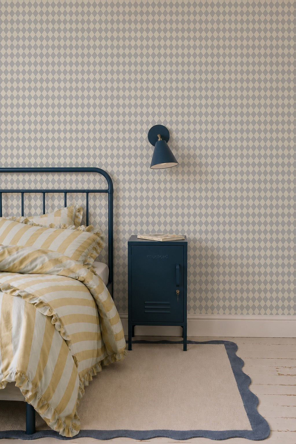 A neatly made bed with yellow and white striped bedding sits beside a blue metal nightstand and lamp, set against Diamond Dancer Wallpaper in Blue, with a light rug on wooden floors.