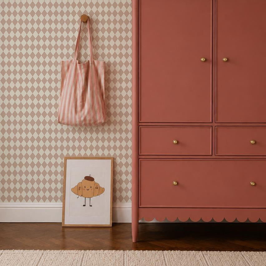 A pink dresser stands on a wooden floor beside a framed croissant drawing, a striped pink bag on a wall hook, and Diamond Dancer Wallpaper in Rose with soft beige harlequin diamond patterns in the background.