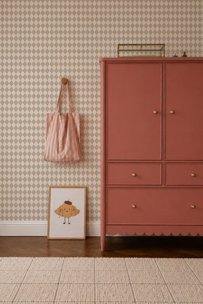 A pink dresser stands on a wooden floor beside a framed croissant drawing, a striped pink bag on a wall hook, and Diamond Dancer Wallpaper in Rose with soft beige harlequin diamond patterns in the background.