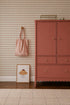 A pink dresser stands on a wooden floor beside a framed croissant drawing, a striped pink bag on a wall hook, and Diamond Dancer Wallpaper in Rose with soft beige harlequin diamond patterns in the background.