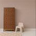 A brown wooden dresser with six drawers stands next to a small beige plastic chair. The background features Diamond Dancer Wallpaper in the Stone Rose colorway, showcasing retro harlequin patterns in shades of brown and cream. A beige rug lies on the light wood floor.