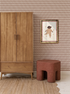 A cozy room with Diamond Dancer Wallpaper in Stone Rose, a wooden wardrobe, a textured brown stool, a rug, and a framed picture of a bear in a clown outfit on the wall.