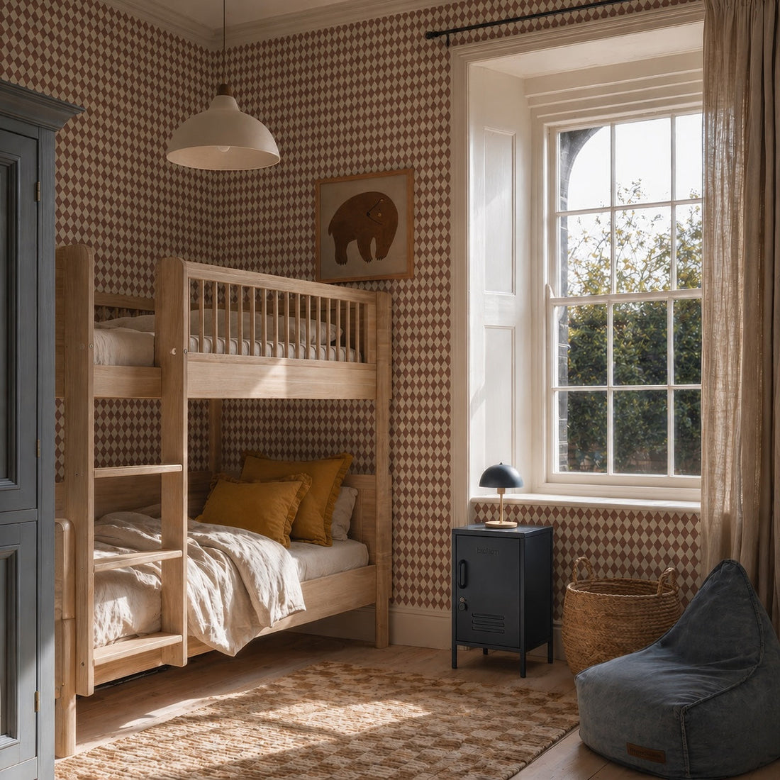 A cozy bedroom with a wooden bunk bed, neutral bedding, Diamond Dancer Wallpaper in Stone Rose, a window with beige curtains, a small black nightstand, bean bag chair, wicker basket, and framed elephant picture on the wall.