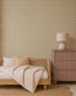 A cozy bedroom with a wooden bed, beige-striped bedding and pillows, a pinkish-brown dresser topped with a round lamp, a scalloped jute rug, and striking Diamond Dancer Wallpaper in yellow.