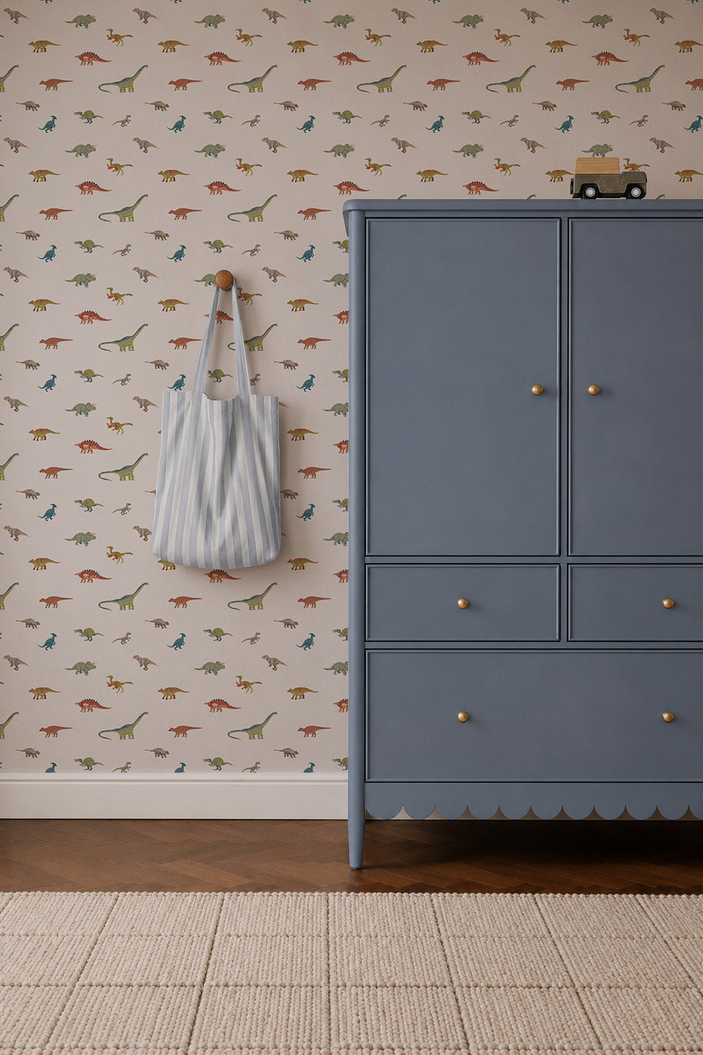 A blue dresser sits on a brown floor by a wall covered with Dino Discovery Wallpaper. A striped tote hangs from a peg for little explorers, while a toy car is placed on the dresser.
