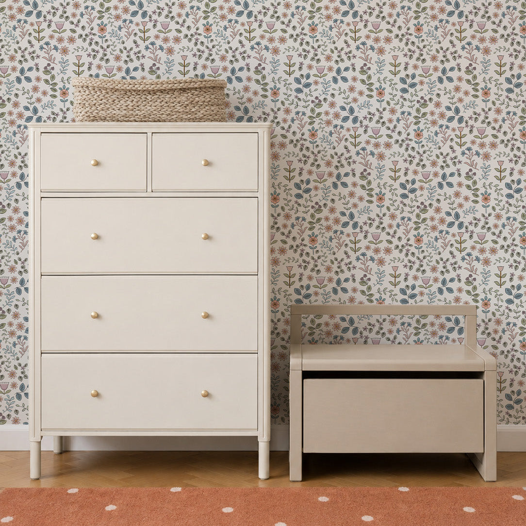 A white dresser and a small beige bench sit on a wooden floor with a peach polka-dot rug. The wall behind features Ditsy Blooms Wallpaper adorned with colorful small flowers. A wicker basket rests on top of the dresser.
