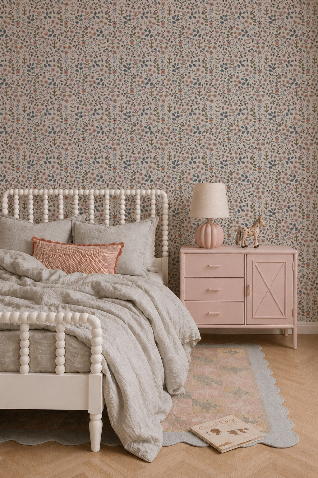 A cozy bedroom featuring a white spindle bed with beige bedding, a pink nightstand displaying a lamp and animal figurines, charming Ditsy Blooms Wallpaper adorning the walls, and a book and rug placed on the wooden floor.