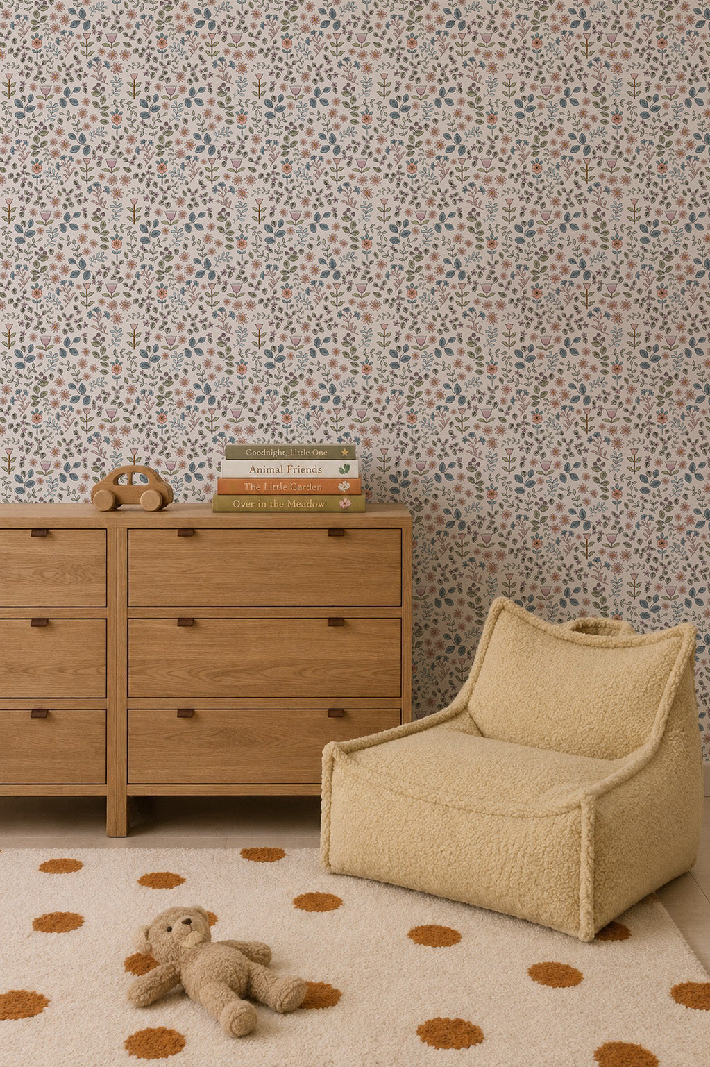 A cozy nursery featuring a wooden dresser, a beige lounge chair, a stuffed bear on a polka dot rug, stacked children’s books, and the charming Ditsy Blooms Wallpaper.
