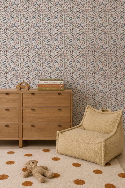 A cozy nursery featuring a wooden dresser, a beige lounge chair, a stuffed bear on a polka dot rug, stacked children’s books, and the charming Ditsy Blooms Wallpaper.