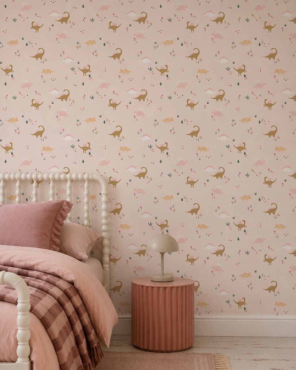 A cozy bedroom with a bed in pink linens, a round pink nightstand with a beige lamp, and playful Dixie Dinosaur Wallpaper featuring deer and florals on a light pink background.