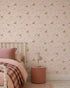 A cozy bedroom with a bed in pink linens, a round pink nightstand with a beige lamp, and playful Dixie Dinosaur Wallpaper featuring deer and florals on a light pink background.