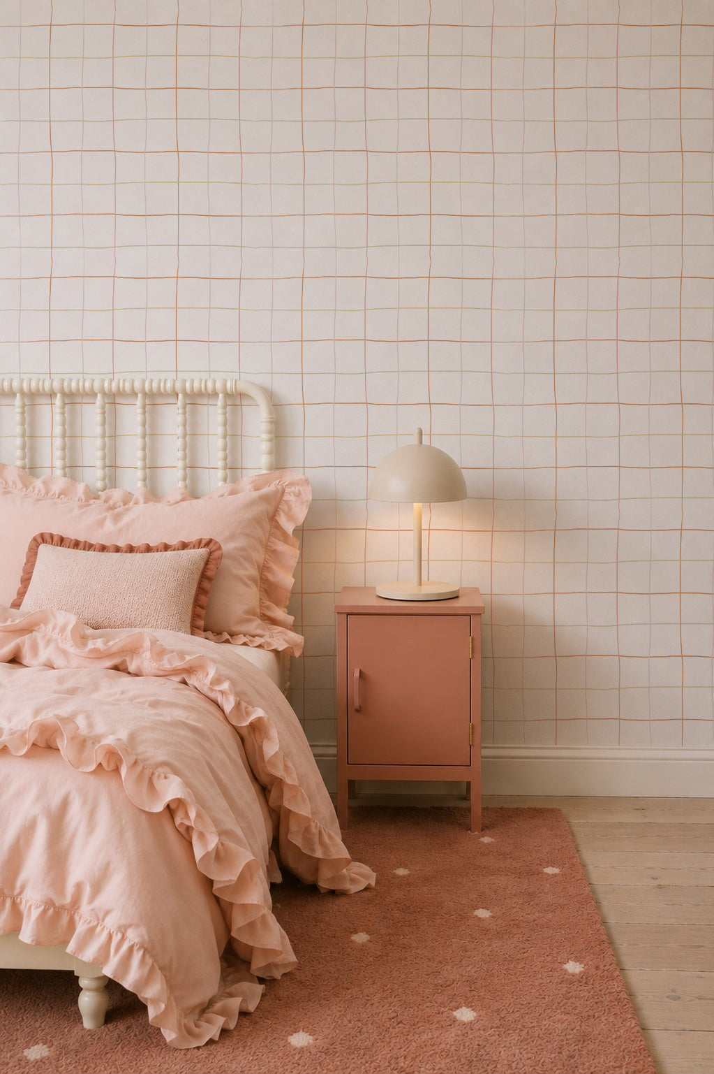 A cozy bedroom features modern interiors with a cream bed frame, blush pink ruffled bedding, a matching pink nightstand, round beige lamp, pink rug with white dots, and Doodle Grid Wallpaper on the white wall.