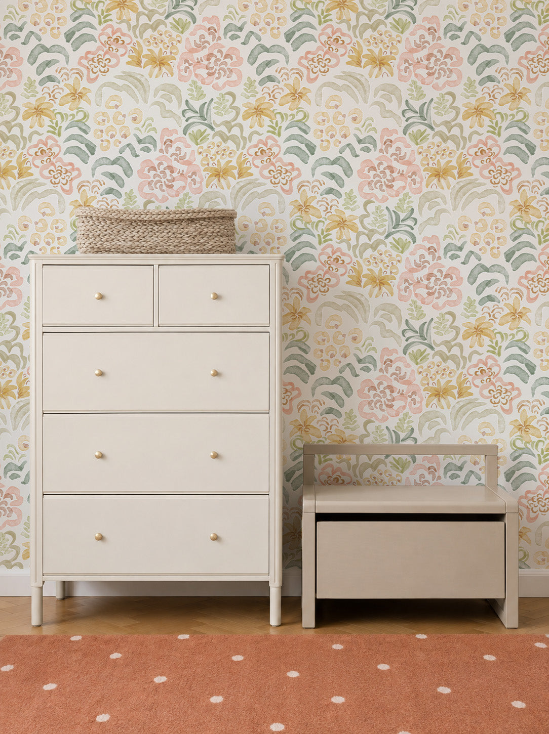 A light-colored dresser and a small storage bench sit against a wall adorned with Floret Dreamscape Wallpaper. A woven basket is on the dresser, and the floor features a rust-colored rug with white polka dots.