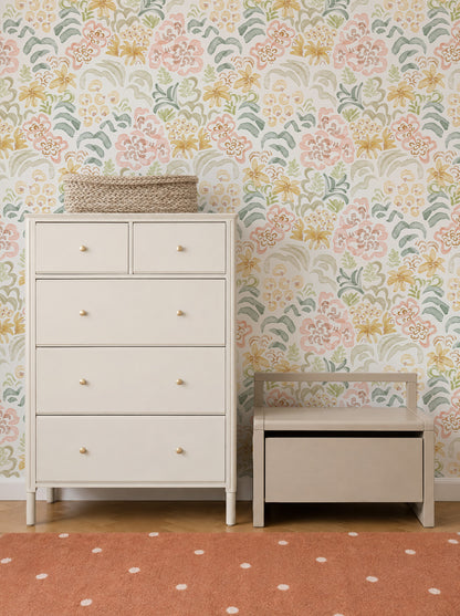 A light-colored dresser and a small storage bench sit against a wall adorned with Floret Dreamscape Wallpaper. A woven basket is on the dresser, and the floor features a rust-colored rug with white polka dots.