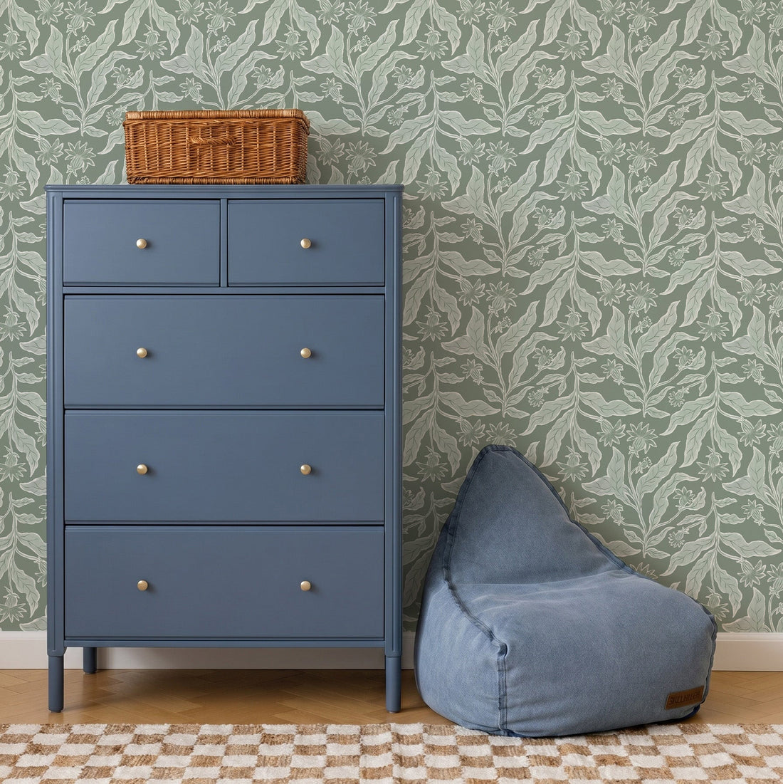 A blue chest of drawers with silver knobs stands by a blue bean bag chair on a checkered rug. A wicker basket sits atop the dresser. The backdrop features Elodies Thistle Wallpaper in sage with green leaf patterns.