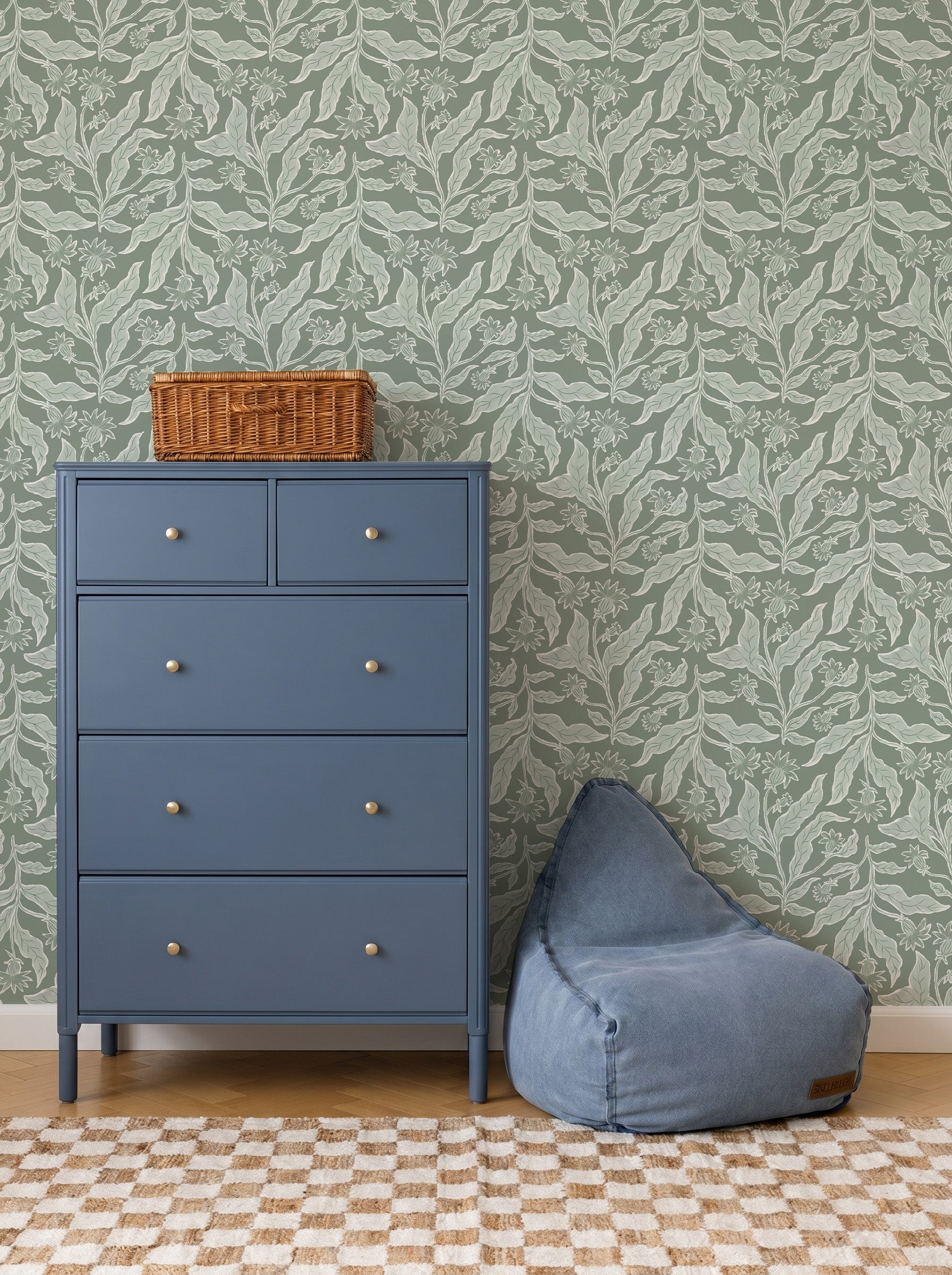 A blue chest of drawers with silver knobs stands by a blue bean bag chair on a checkered rug. A wicker basket sits atop the dresser. The backdrop features Elodies Thistle Wallpaper in sage with green leaf patterns.