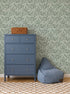 A blue chest of drawers with silver knobs stands by a blue bean bag chair on a checkered rug. A wicker basket sits atop the dresser. The backdrop features Elodies Thistle Wallpaper in sage with green leaf patterns.