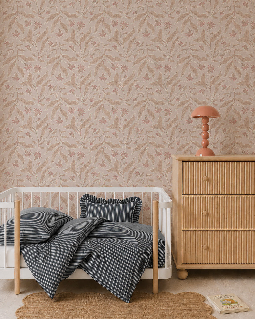 A cozy nursery with whimsical decor features a small white crib, blue-striped bedding, a round rug, and a wooden dresser with a pink lamp. Elodies Thistle Wallpaper in Sand adds charm in soft beige and pink tones, while a book rests near the dresser.