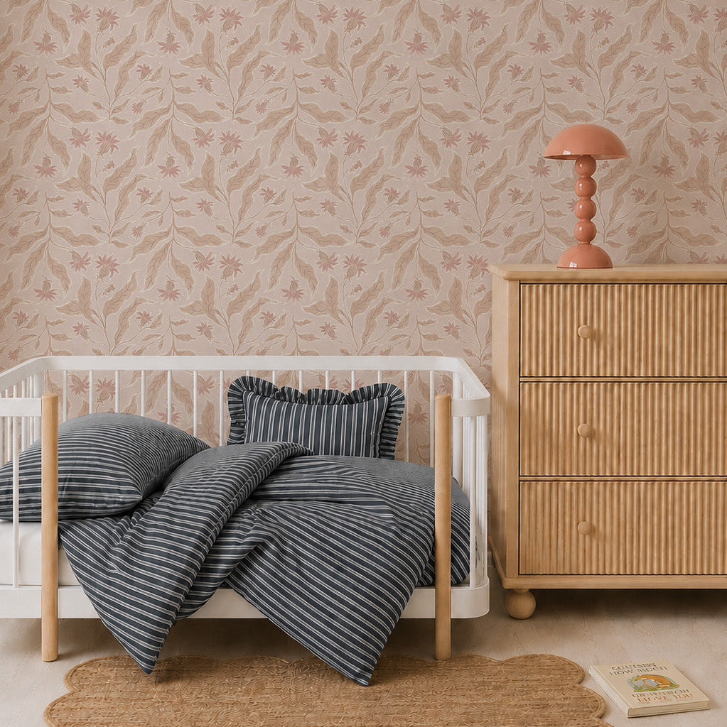 A cozy nursery with whimsical decor features a small white crib, blue-striped bedding, a round rug, and a wooden dresser with a pink lamp. Elodies Thistle Wallpaper in Sand adds charm in soft beige and pink tones, while a book rests near the dresser.