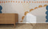 A wooden dresser stands on a woven rug before a wall adorned with the Evie Abstract Wallpaper Mural, featuring blue, mustard, and beige dots, triangles, scallops, and lines. Sunlight highlights this colorful setting.