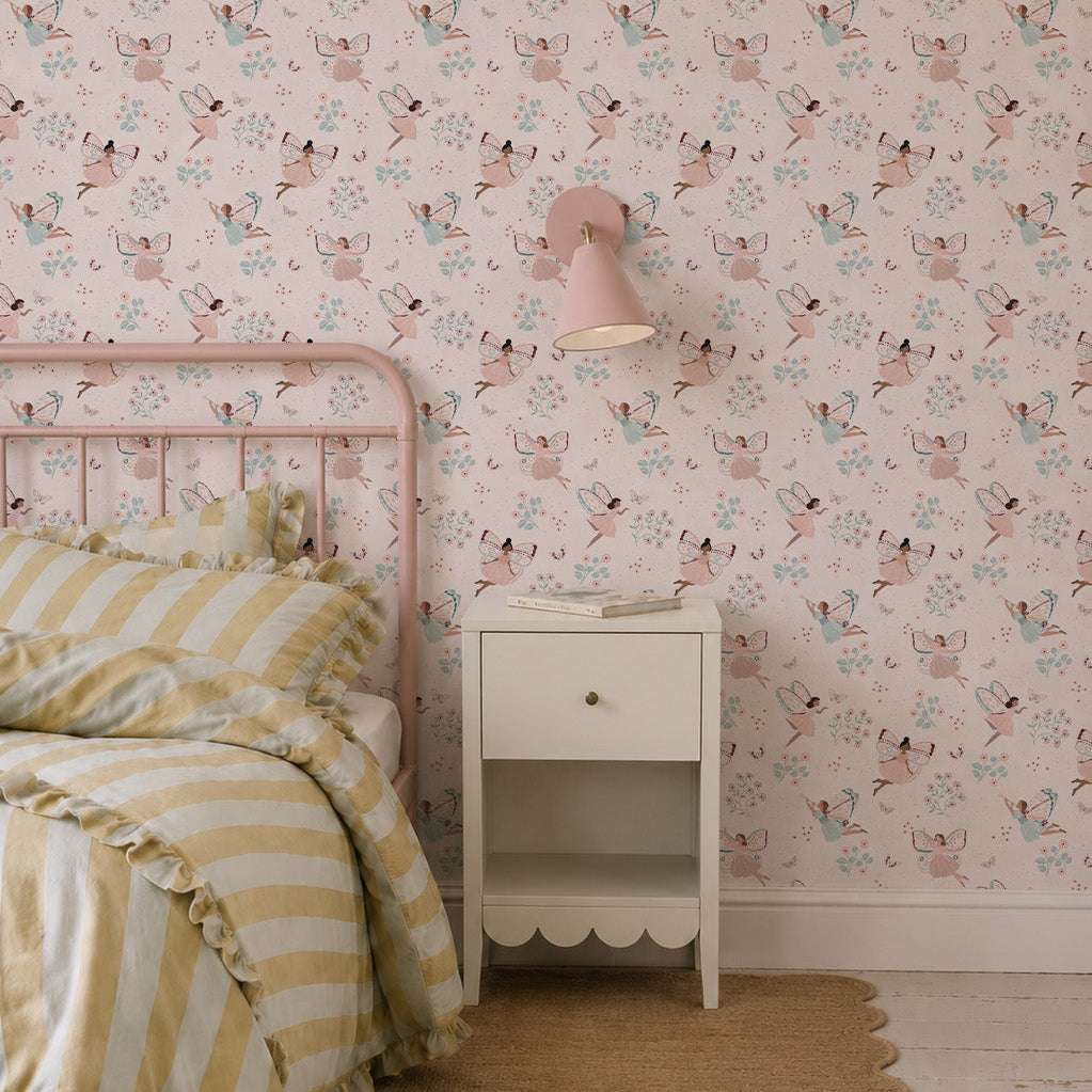 A cozy bedroom with a pink metal bed, yellow-striped bedding, white nightstand, pink wall lamp, and Fairy Wonderland Pinks Wallpaper featuring whimsical birds and flowers. A woven rug lies on the wooden floor.