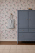 A blue dresser with gold knobs sits on a tan rug. A gray and white striped bag hangs on the wall, which is decorated with Fairy Wonderland Purple Wallpaper featuring playful floral designs.