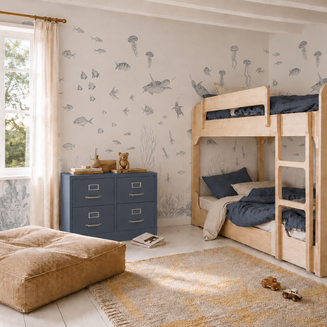 Children’s bedroom with wooden bunk beds, a blue dresser, tan pouf, and a rug. The walls feature the Fish Tank Mural in blue, filled with sea creatures like fish, jellyfish, and underwater plants. Sunlight streams in from a window with beige curtains.