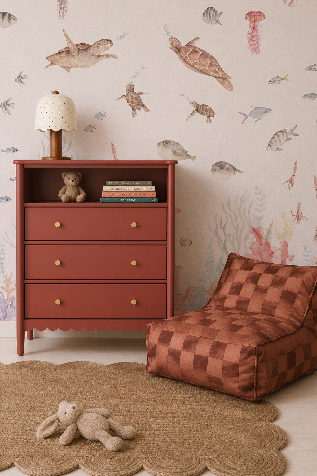 A cozy nursery includes a rust-colored dresser with books and a teddy bear, a checkered pouf chair, a round woven rug with a plush bunny, and an ocean-themed wall featuring the Fish Tank Mural.