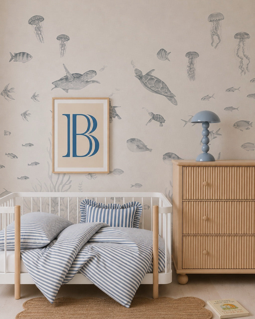 A cozy child’s bedroom with a Fish Tank Mural, Blue featuring playful sea creatures, a small wooden bed with blue striped bedding, a dresser with a blue lamp, a framed letter “B,” and a scalloped jute rug on a light floor.