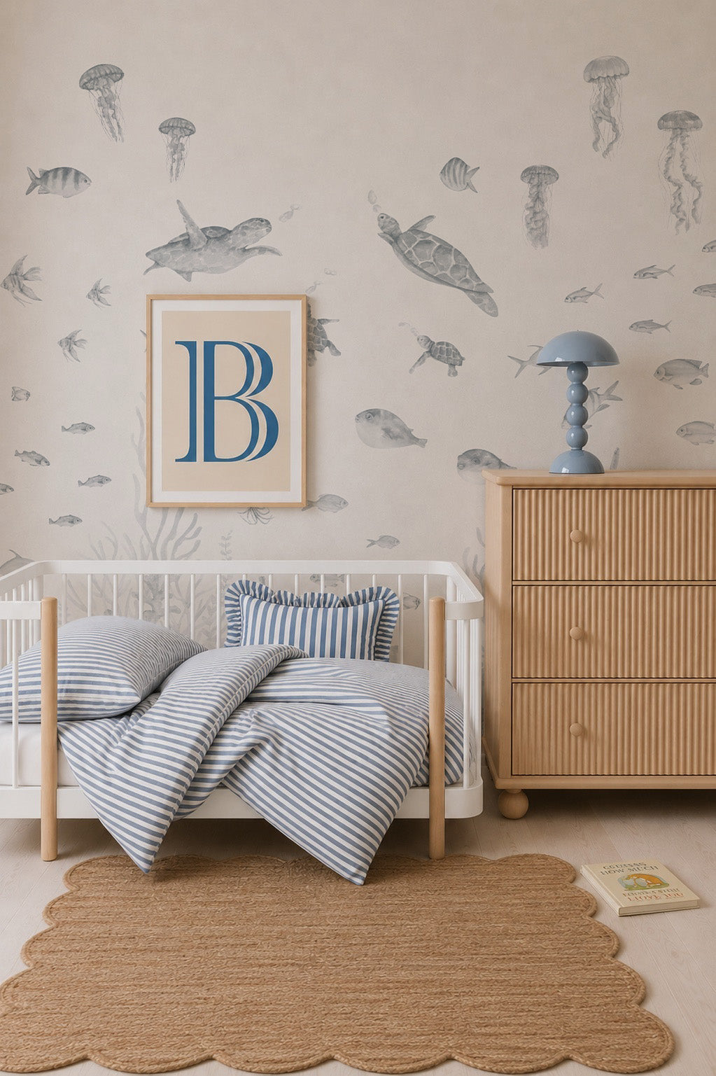A nursery with a wooden crib, blue striped bedding, a rattan dresser, a blue lamp, and the Fish Tank Mural in Blue featuring playful sea creatures. A framed letter B hangs above the crib, and a scalloped rug is on the floor.