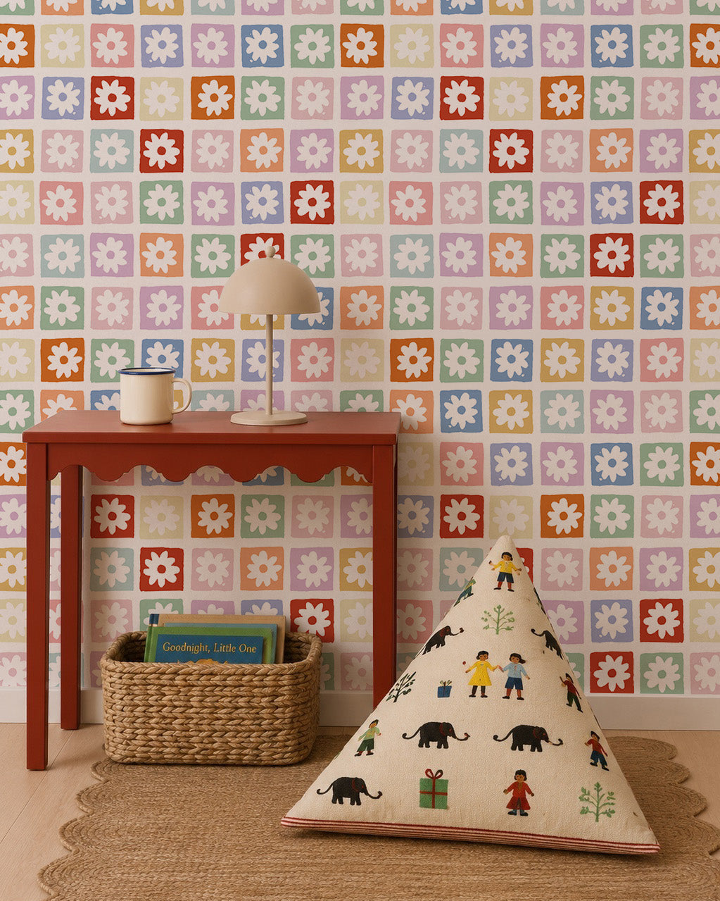 A colorful room features a red table with a lamp and cup, a woven basket with books underneath, a patterned floor cushion, and walls adorned in the Floral Funland Mural showcasing multicolored floral squares.