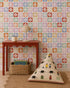 A colorful room features a red table with a lamp and cup, a woven basket with books underneath, a patterned floor cushion, and walls adorned in the Floral Funland Mural showcasing multicolored floral squares.