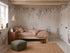 A cozy bedroom features the Florence Florals Mural as wallpaper, a wooden bed with pink bedding, elegant nursery decor, a brown nightstand with books and plants, a woven rug, green pouf, teddy bear, and a pendant light from the beamed ceiling.