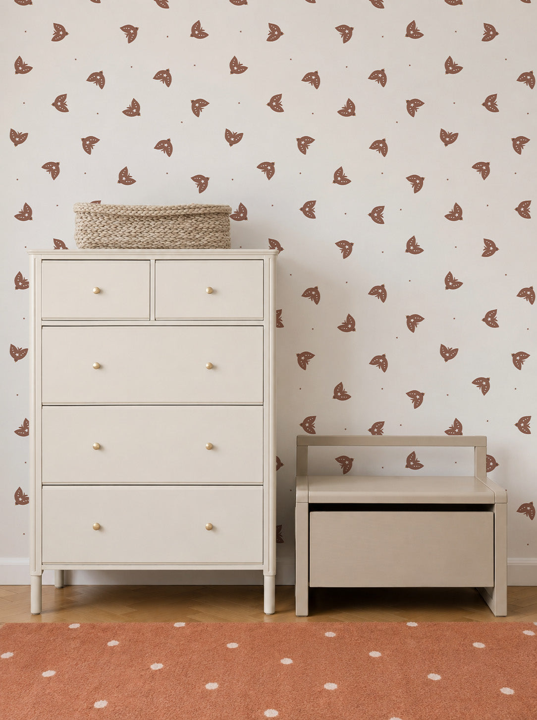 A cream-colored dresser and a small bench sit against a wall adorned with Flutter Sky Wallpaper Mocha in soft brown tones. A woven basket rests on the dresser, and an orange rug with white dots covers the wooden floor.