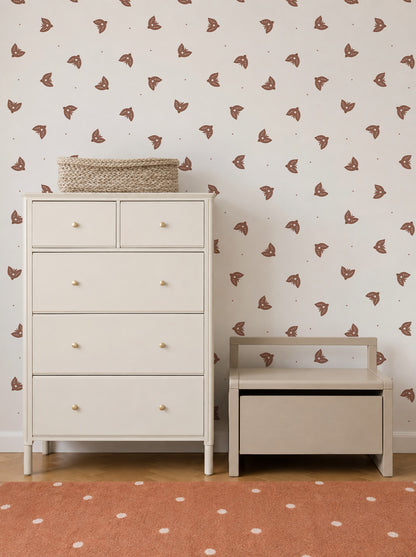 A cream-colored dresser and a small bench sit against a wall adorned with Flutter Sky Wallpaper Mocha in soft brown tones. A woven basket rests on the dresser, and an orange rug with white dots covers the wooden floor.
