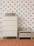 A cream-colored dresser and a small bench sit against a wall adorned with Flutter Sky Wallpaper Mocha in soft brown tones. A woven basket rests on the dresser, and an orange rug with white dots covers the wooden floor.