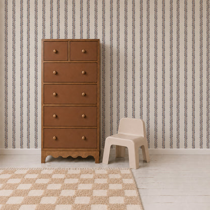 A brown wooden dresser stands next to a small beige plastic chair on a white floor, with a checkered rug and Folk Scallop Stripe Wallpaper in Chambray &amp; Mocha in the background.