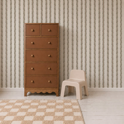 A tall brown dresser with six drawers stands next to a small beige plastic chair, on a white wood floor with a beige checkered rug and Folk Scallop Stripe Wallpaper in Olive &amp; Mocha in the background.