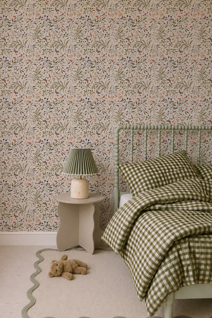 A cozy bedroom with a green plaid bed, a scalloped nightstand holding a lamp, a teddy bear on a cream rug with wavy edges, and the wall adorned in Forest Floor Wallpaper featuring delicate floral patterns.