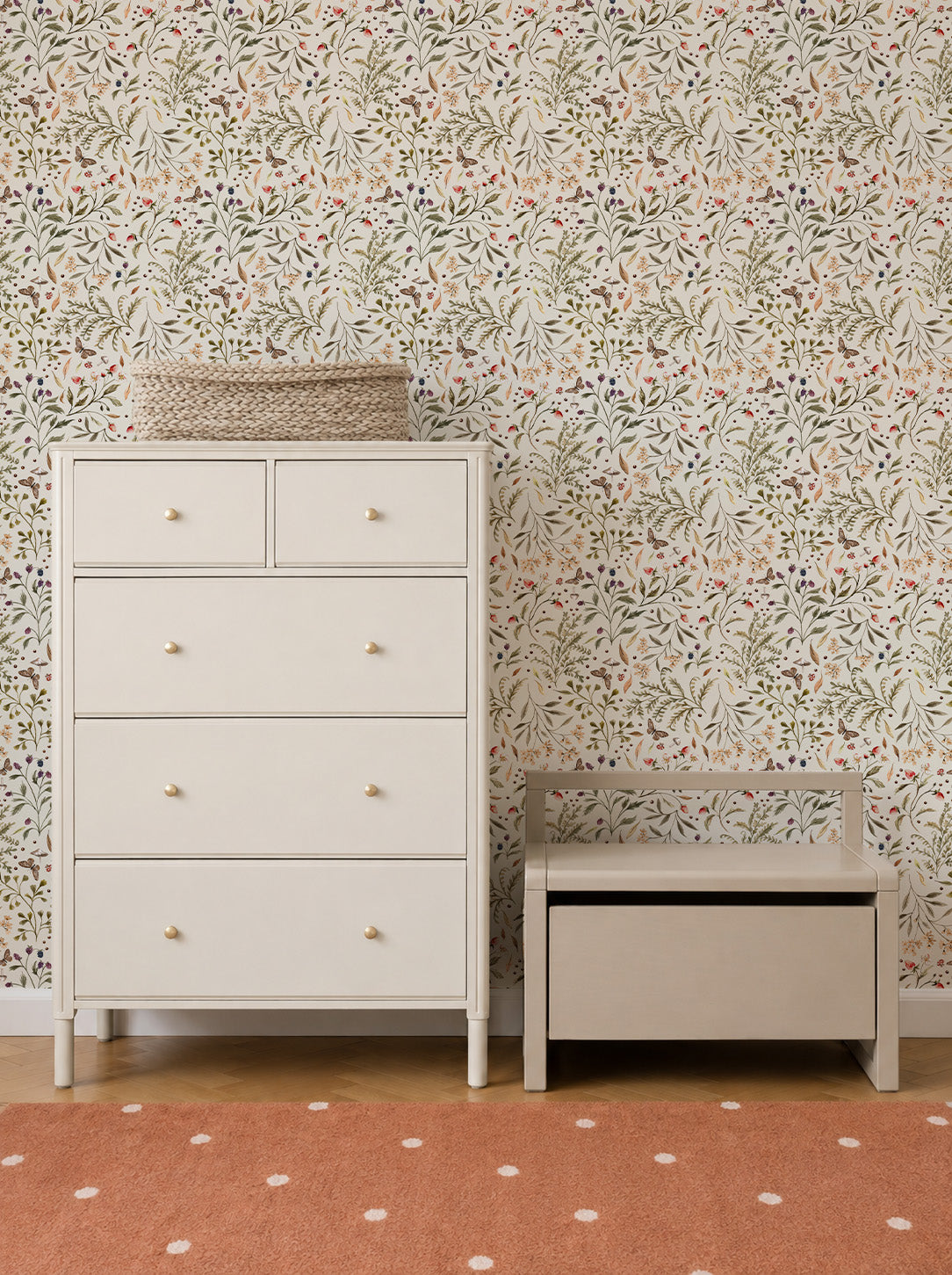 White dresser with two small and three large drawers beside a small storage bench. A woven basket sits on the dresser. The background features Forest Floor Wallpaper with floral motifs, and an orange rug with white dots is on the floor.