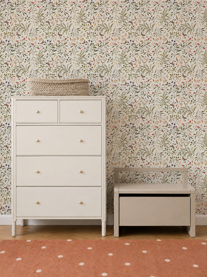 White dresser with two small and three large drawers beside a small storage bench. A woven basket sits on the dresser. The background features Forest Floor Wallpaper with floral motifs, and an orange rug with white dots is on the floor.
