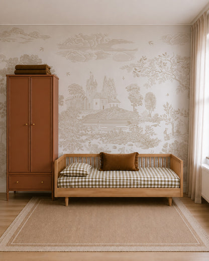 A cozy bedroom with a wooden daybed, plaid bedding, a rust-colored pillow, a tall rust wardrobe, and beige nature wallpaper—the Forest Hideaway Mural in Neutral—featuring a scenic countryside illustration with a castle and trees. Light streams in through a sheer-curtained window.