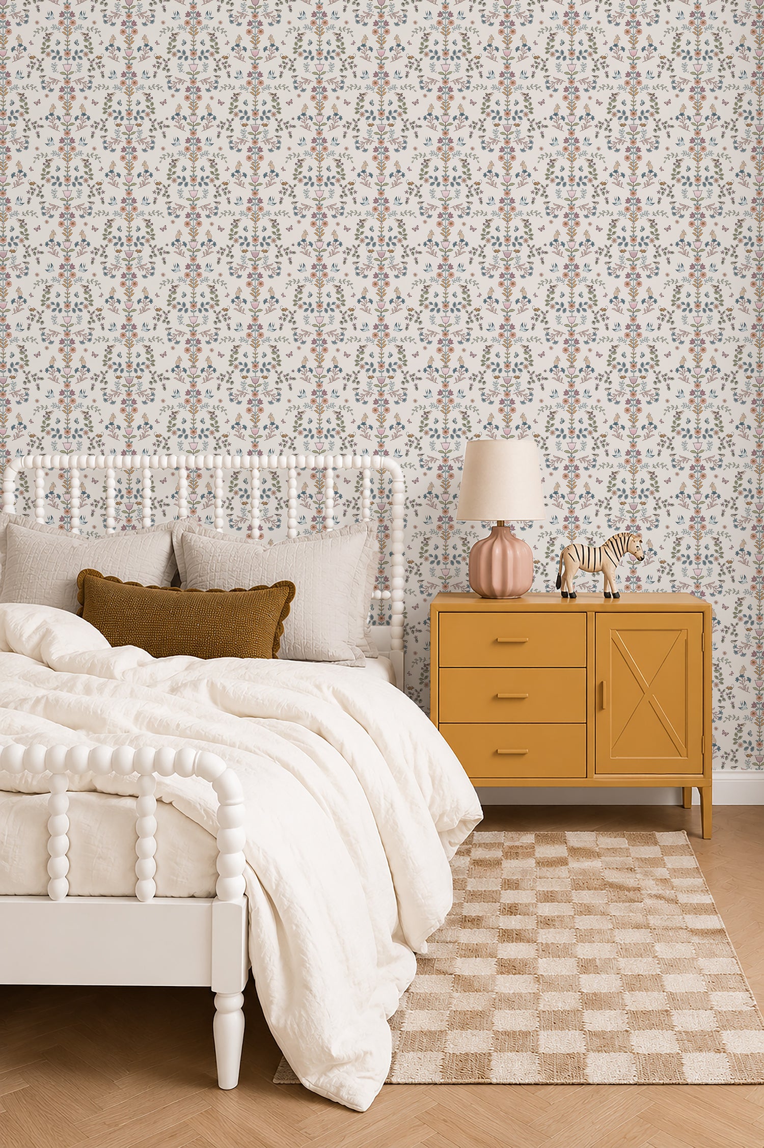 A cozy bedroom features a white spindle bed, cream bedding, textured pillows, a mustard yellow nightstand, pink lamp, zebra figurine, Forest Tales Wallpaper, and a checkered rug on a wooden floor.