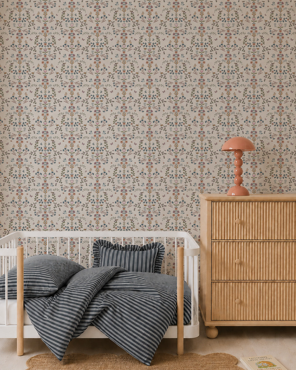 A cozy nursery features a rattan dresser, a white crib with blue-striped bedding, a scalloped jute rug, and Forest Tales Wallpaper. A pink mushroom-shaped lamp sits on the dresser, with a book on the floor nearby.