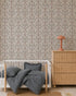 A cozy nursery features a rattan dresser, a white crib with blue-striped bedding, a scalloped jute rug, and Forest Tales Wallpaper. A pink mushroom-shaped lamp sits on the dresser, with a book on the floor nearby.