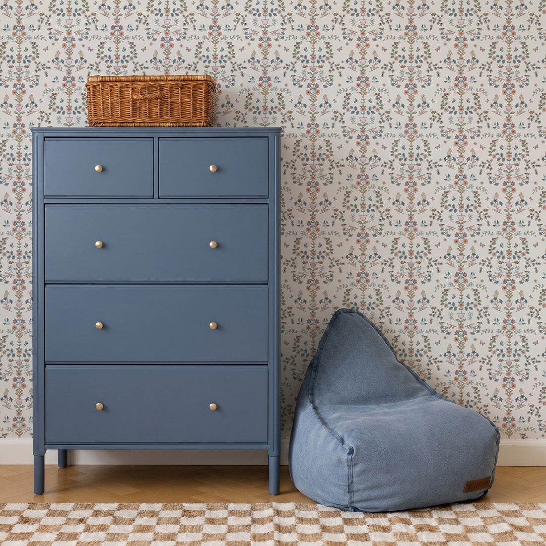 A blue dresser with silver knobs stands next to a blue bean bag chair. A wicker basket sits on the dresser, while Forest Tales Wallpaper with whimsical floral patterns decorates the background above a checkered rug on a wooden floor.